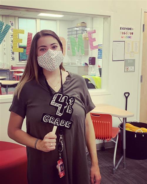 Ms. Hewey wearing a mask standing in front of her classroom. She is holding a sign that says 4th grade. Ms. Hewey wearing a mask standing in front of her classroom. She is holding a sign that says 4th grade.