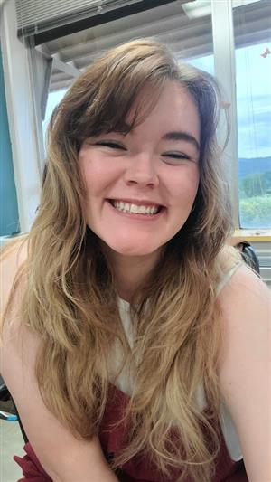Hayley is sitting and smiling- long blonde hair with bangs, red jumpsuit, and white tank top. 