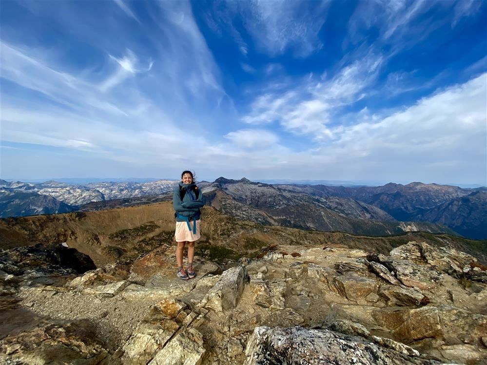 woman stands on a rocky mountain summit with blue sky smattered with clouds in the background
